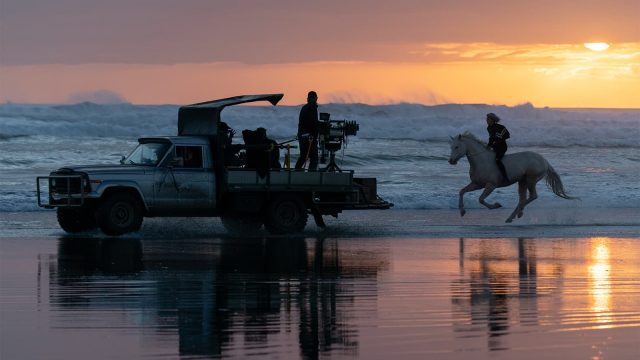 Film crew in a pick up filming a man on a white horse running along the sea edge